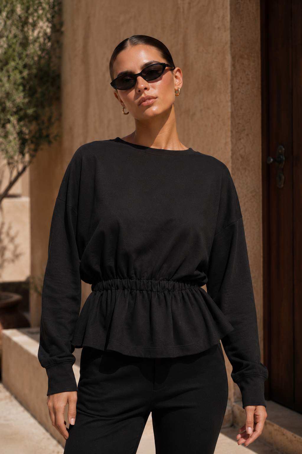 A woman wearing a black long-sleeve top with a gathered peplum waist and black pants, standing outdoors against a beige stucco wall and dark wooden door.